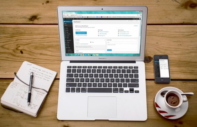 Portátil com o painel do WordPress aberto, junto a um caderno, um telemóvel e uma chávena de café.