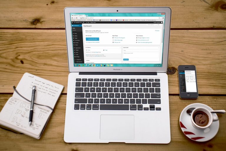 Portátil com o painel do WordPress aberto, junto a um caderno, um telemóvel e uma chávena de café.