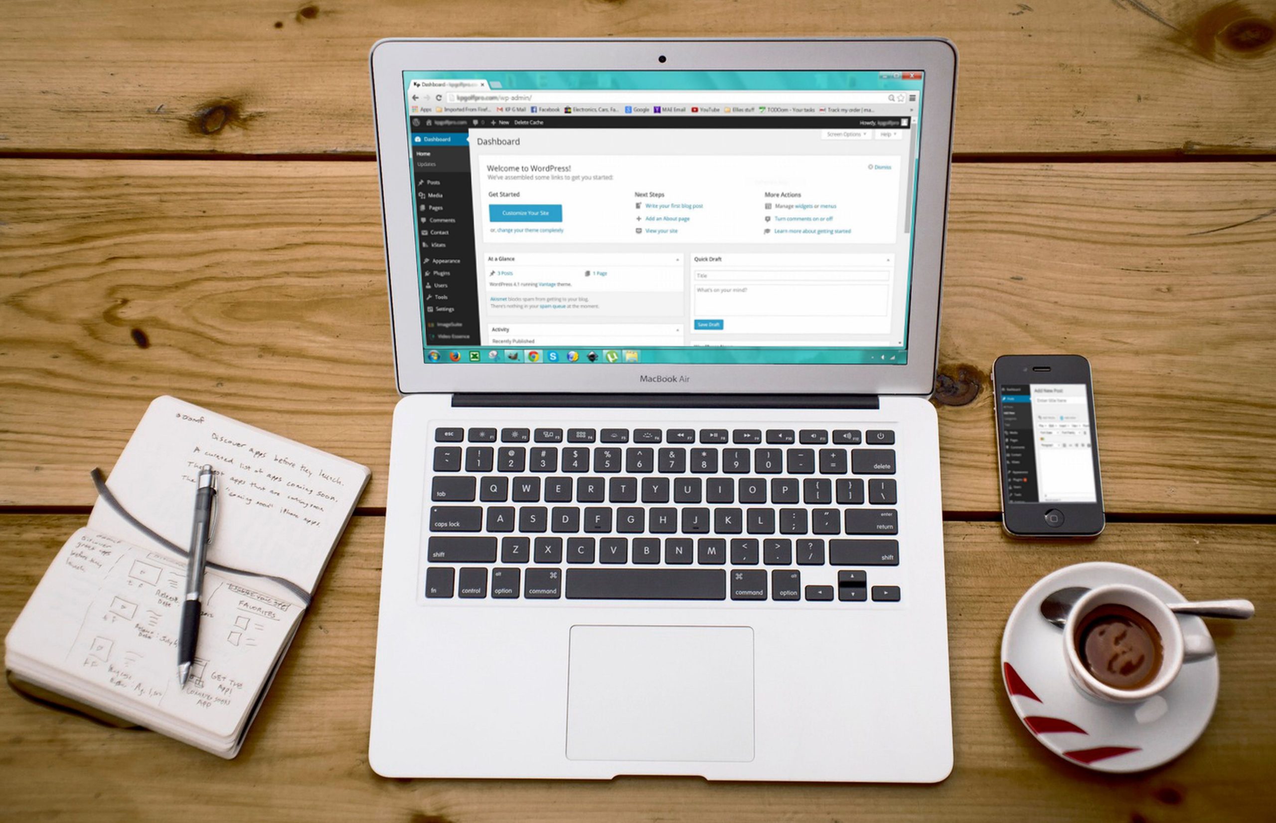 Portátil com o painel do WordPress aberto, junto a um caderno, um telemóvel e uma chávena de café.