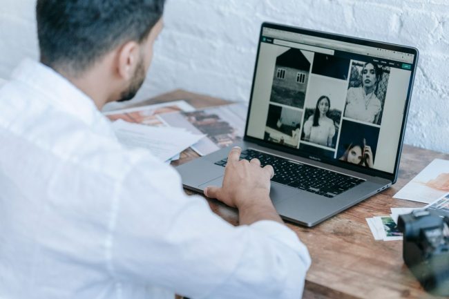 Homem a consultar uma galeria de fotografias num portátil, numa mesa com fotografias impressas e uma câmara.